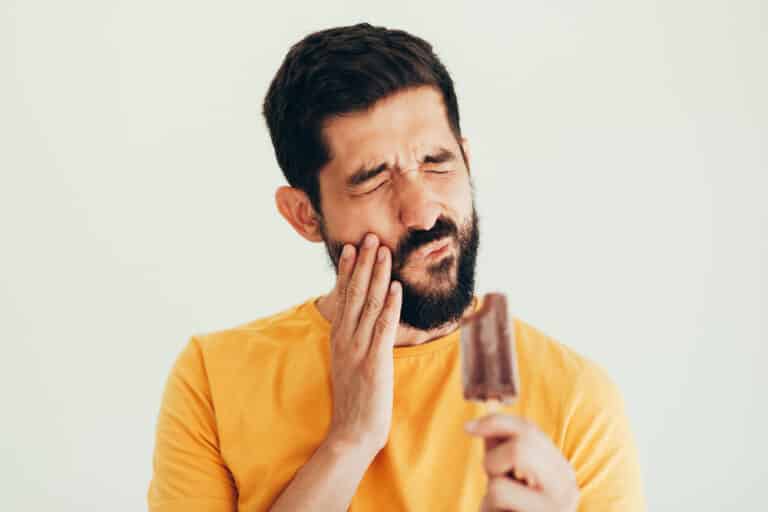 Man wincing and touching hand to cheek due to sensitive teeth, holding ice cream bar